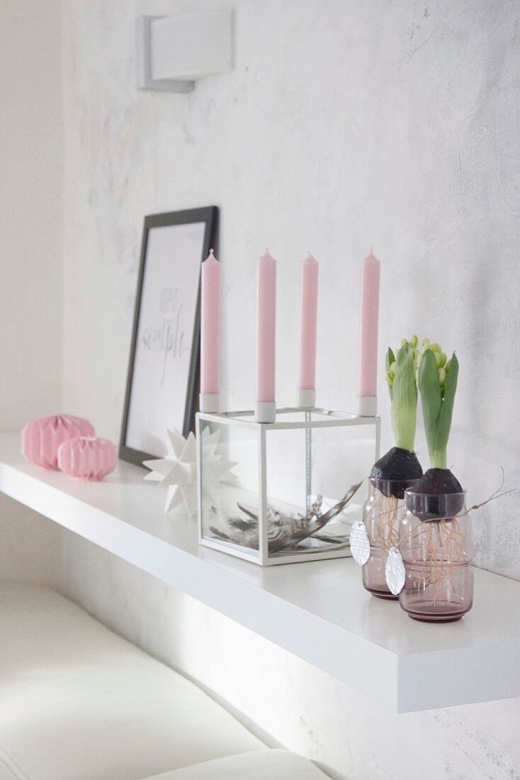 Hyazinthen in Glas, würfelförmiger Kerzenständer mit rosa Kerzen und Papierdeko auf weißer Wandkonsole