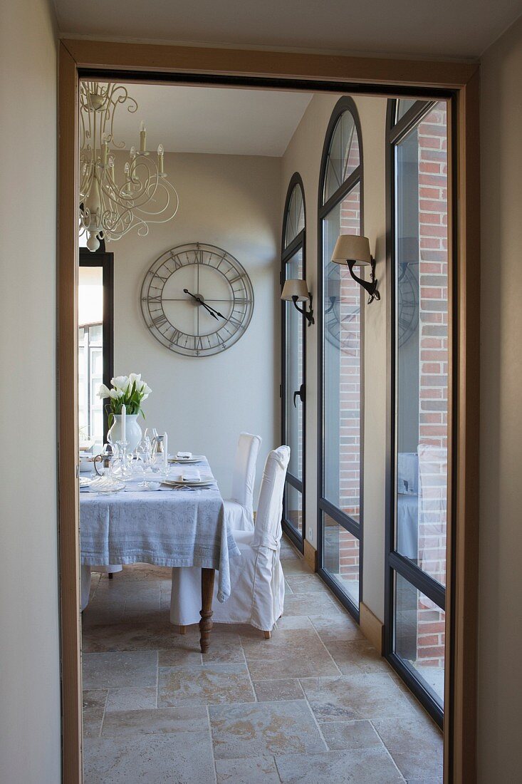 Set dining table and white loose-covered chairs next to floor-to-ceiling arched windows seen through open door