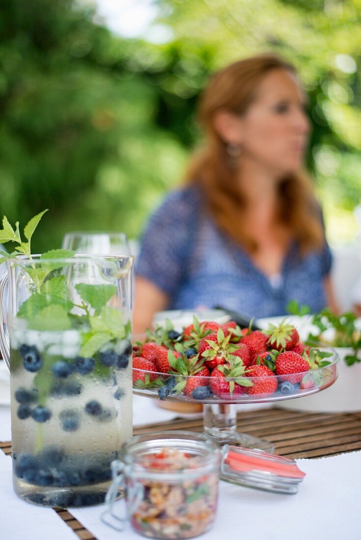 Erfrischungsgetränk mit Blaubeeren in Glaskanne und frische Erdbeeren in Glasschale auf Gartentisch