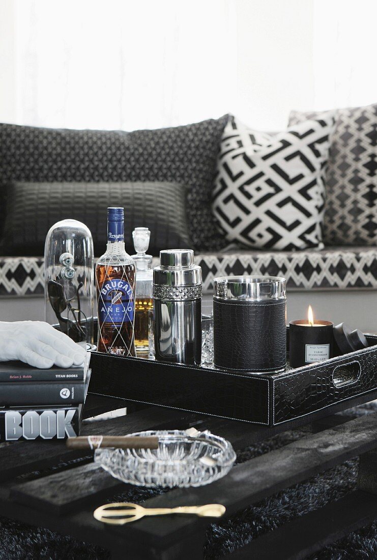 Minibar auf Tablett mit Cocktailzubehör und Aschenbecher mit Zigarre auf schwarzem Palettentisch vor Sofa