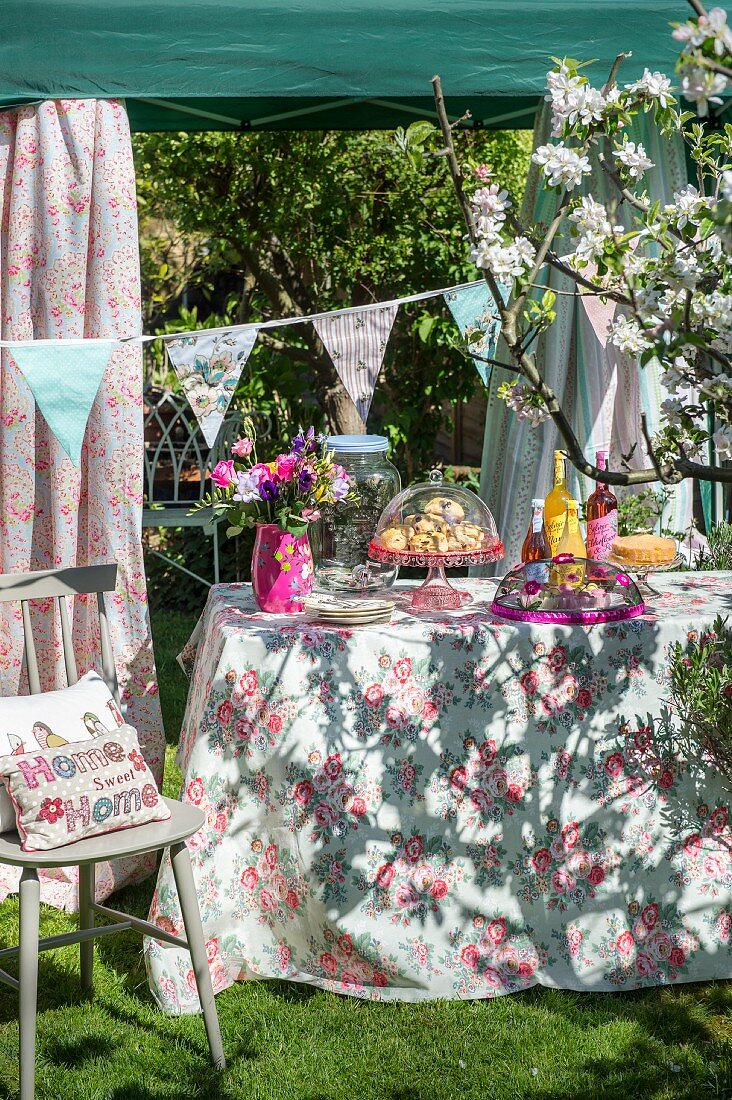 Sonnenbeschienenes Gartenbuffet mit floral gemusterter Tischdecke und Wimpelgirlande vor Pavillon