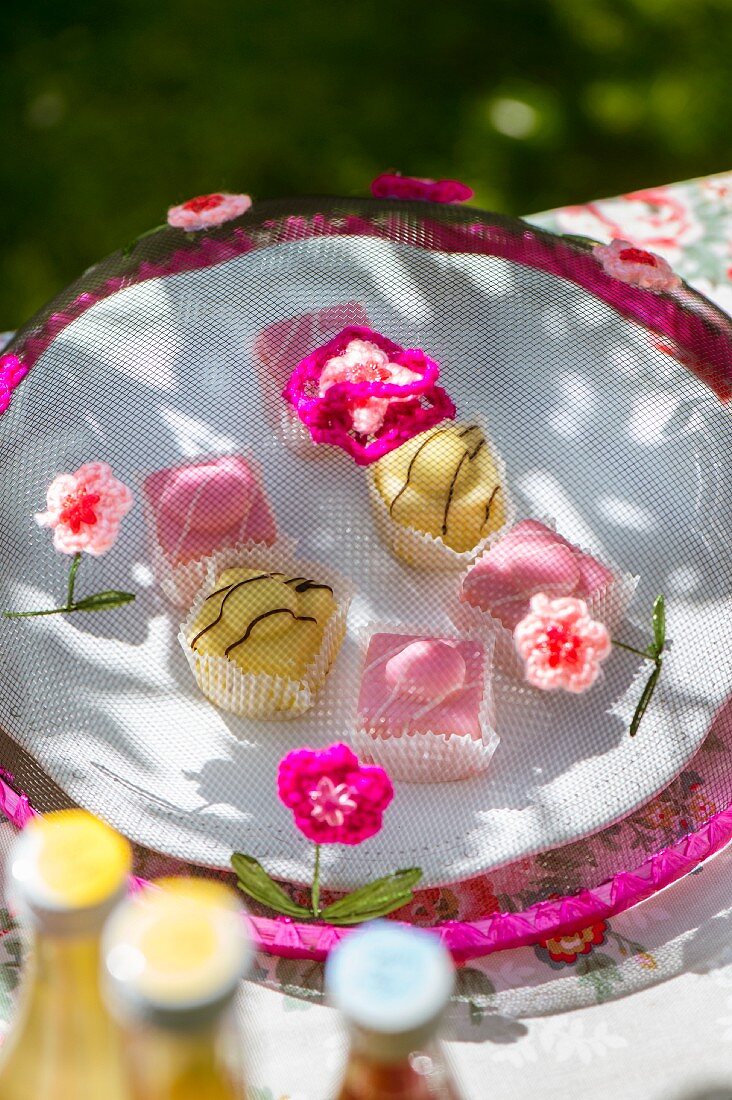 Colourful petit fours under mesh cake cover decorated with crocheted flowers on garden table
