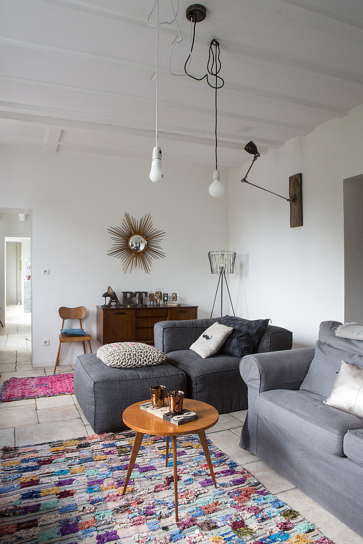 Retro Beistelltisch und graue Polstermöbel in schlichtem Wohnraum, Pendelleuchten mit sichtbaren Leuchtmitteln an weißer Holzbalkendecke