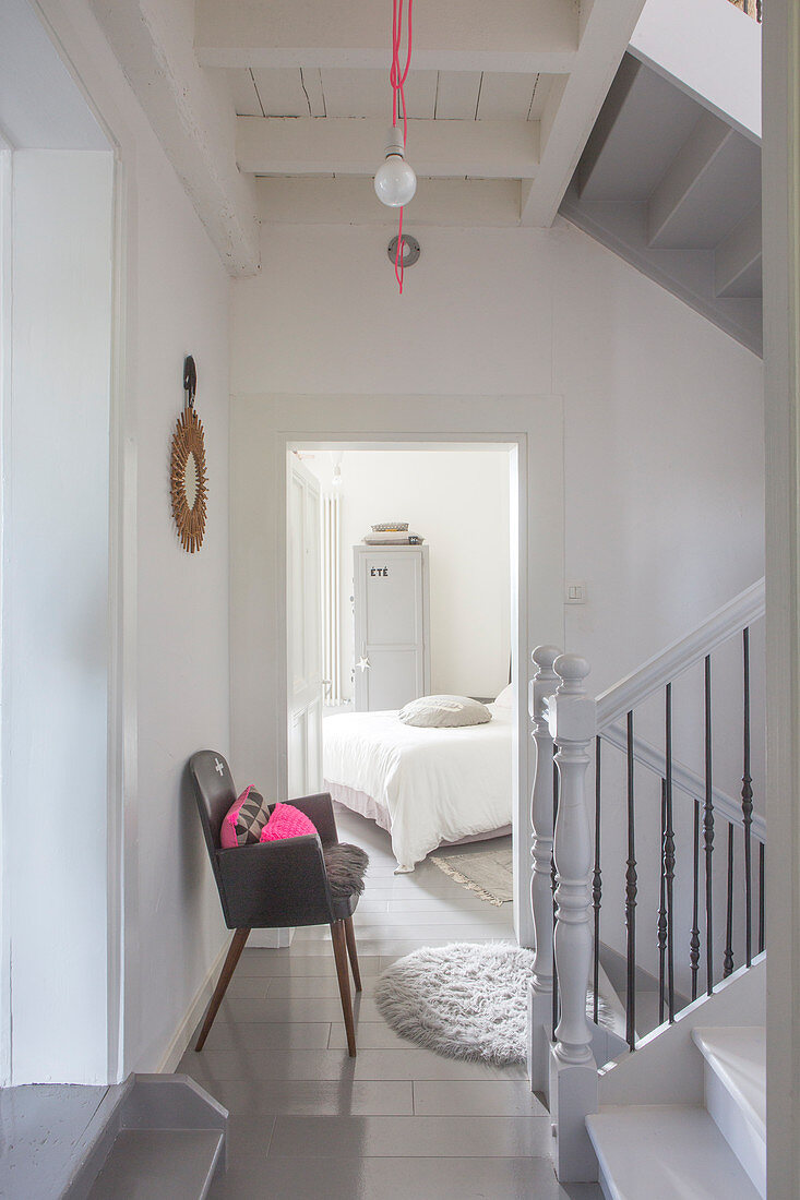 Treppenhaus mit grauem Stuhl vor offener Zimmertür und Blick auf Bett in restauriertem Ambiente
