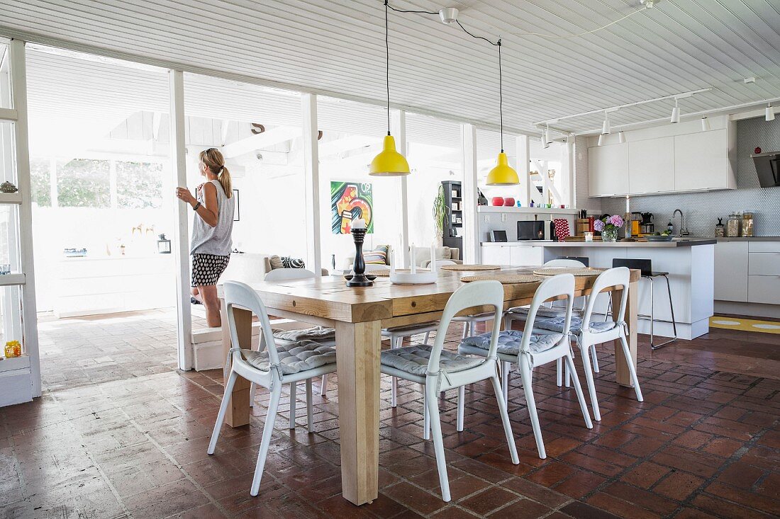 Essplatz mit weissen Stühlen unter gelben Pendelleuchten in offener Küche, Frau vor Fenster