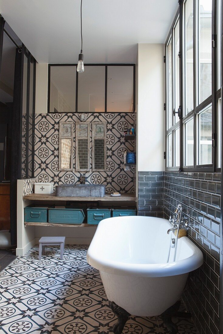 Bad mit Ornamentfliesen, freistehender Badewanne vor Industriefenster und rustikalem Waschtisch mit Metallbehältern auf Ablagebrett