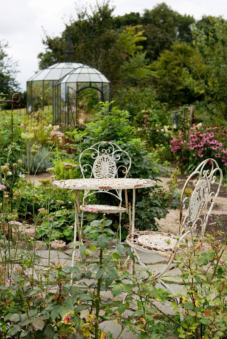Ornate vintage metal chairs and table on stone floor in romantic garden with small orangery in background