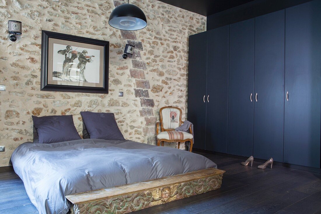 Doppelbett mit graublauer Bettwäsche vor Natursteinwand, seitlich dunkelblauer Kleiderschrank