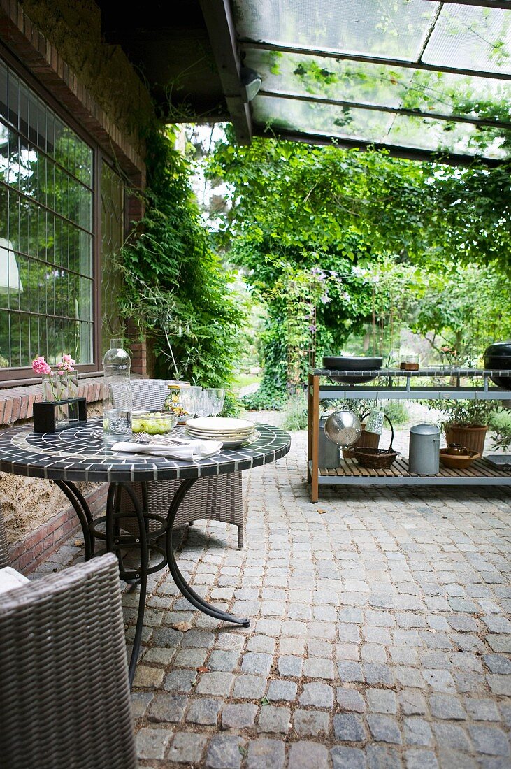 Gartentisch mit Metallgestell und gefliester Tischplatte auf überdachter Terrasse mit Kopfsteinpflaster, im Hintergrund Kochtheke