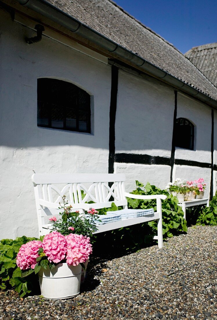 Weiß lackierte Sitzbank mit blühenden Hortensien vor traditionellem Landhaus mit sichtbarem Fachwerk