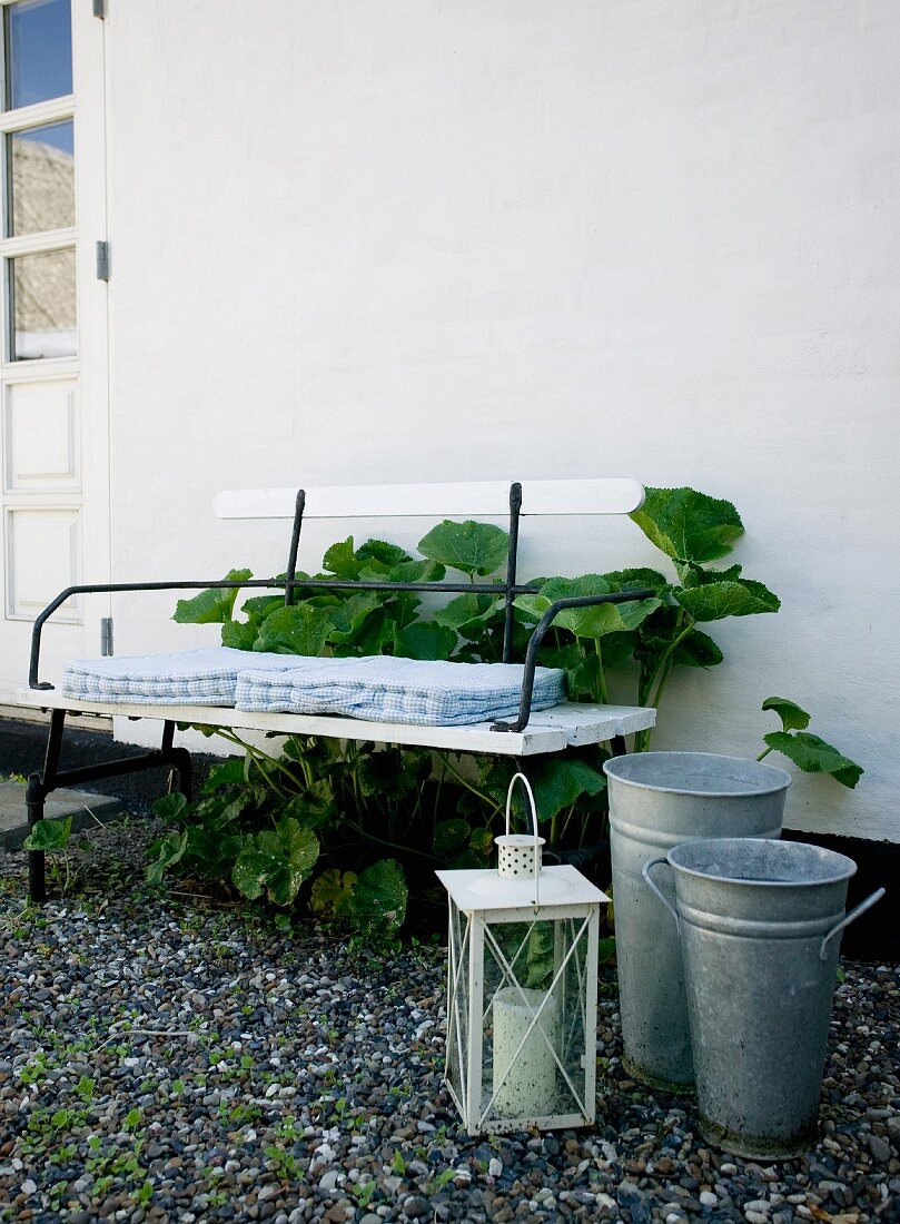 Laterne und zwei Zinkeimer vor weiss lackierter Gartenbank mit schwarzem Metallgestell und Sitzkissen vor Hauswand
