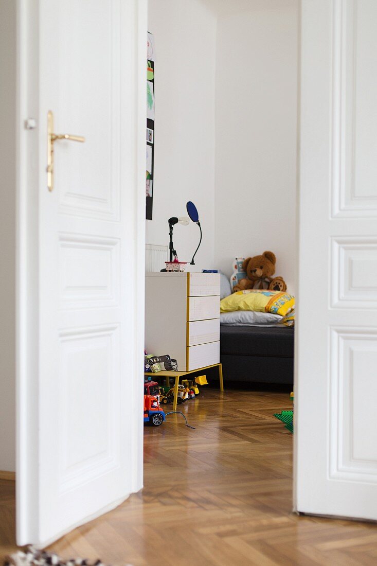 Open white double doors in period apartment with herringbone parquet floor and view into child's bedroom