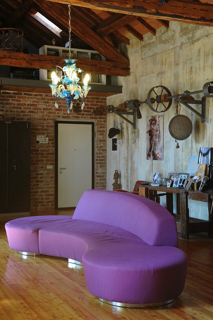 Curved purple designer sofa on wooden floor below glass chandelier in industrial-style loft apartment