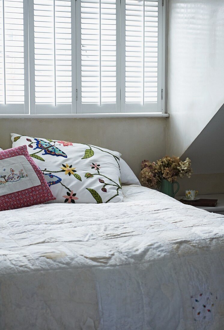 Bett mit heller Tagesdecke und Kissen mit Stickereien vor Fenster mit geschlossenen Innenläden