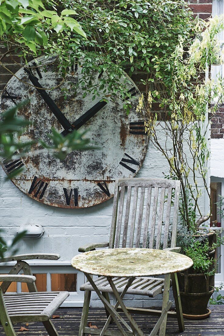 Verwitterter Tisch und Holzstühle auf Terrasse, im Hintergrund Ziffferblatt einer Vintage Bahnhofsuhr an begrünter Ziegelwand