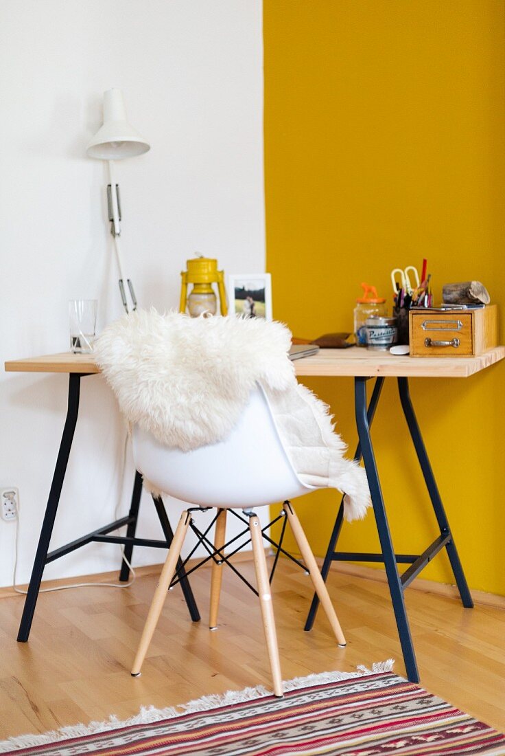 Schreibtisch aus Holzplatte auf Metallgestell in Zimmerecke mit gelber Wand, Schalenstuhl mit weißem Schaffell davor