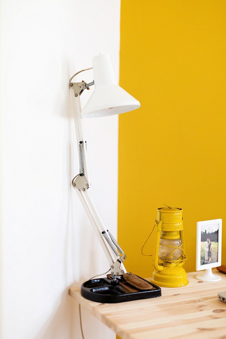 weiße Schreibtischleuchte neben gelber Petroleumlampe auf Holztischplatte in Zimmerecke vor gelber Wand