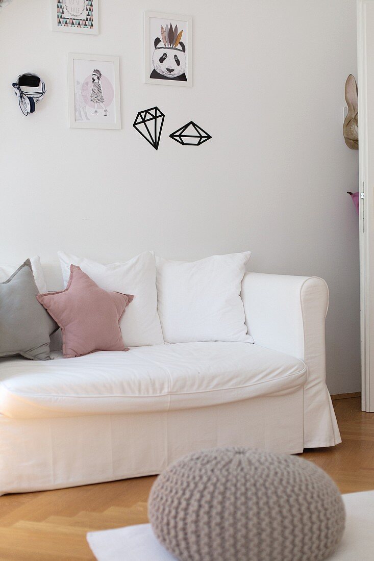 Kissenreihe auf Sofa mit weisser Husse, an Wand gerahmte Zeichnungen und Wandtattoos
