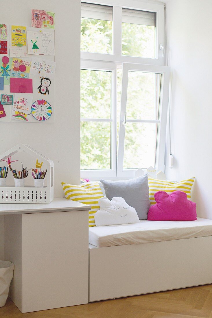 White desk below colourful children's drawings on wall and next to window seat with seat cushion and bright scatter cushions