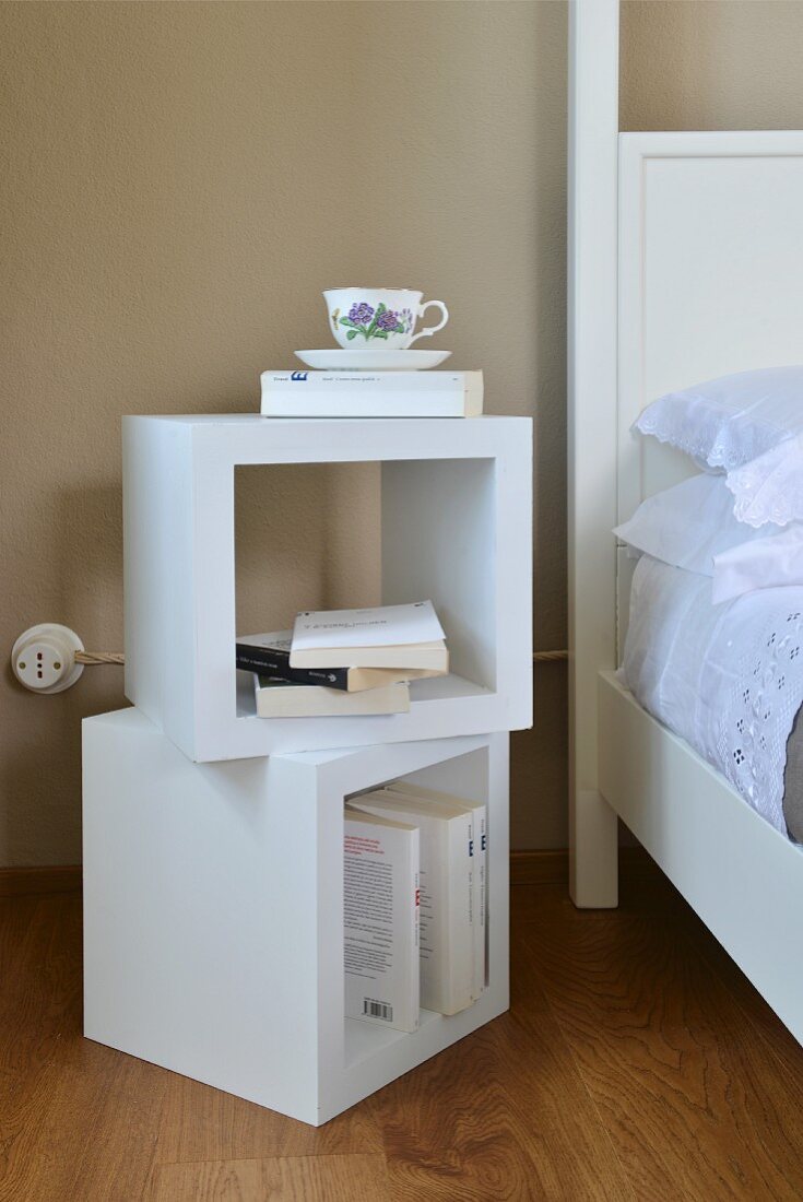 Books and teacup on two stacked cube-shaped shelves next to white bed with lace-trimmed bed linen