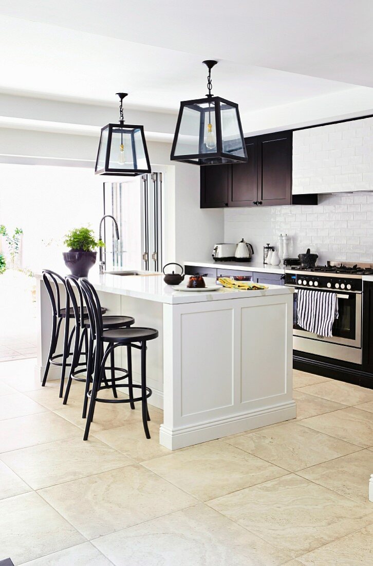 weiße Kücheninsel mit schwarzen Barhockern unter Laternenleuchten vor Küchenzeile und offenen Terrassentüren