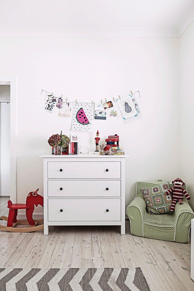Children's room with white chest of drawers, rocking animal and green children's armchair, wall decoration with drawings and photos on a clothesline