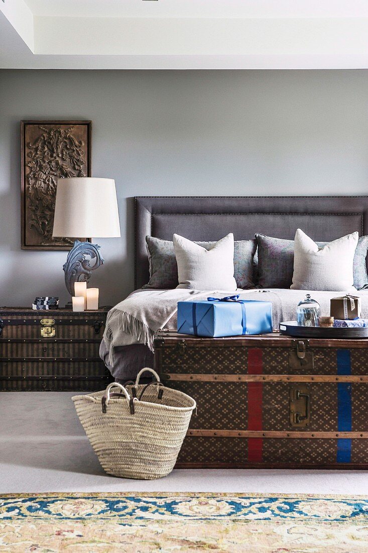 Wrapped gift on antique chest in front of double bed and draped pillows on upholstered headboard in elegant bedroom