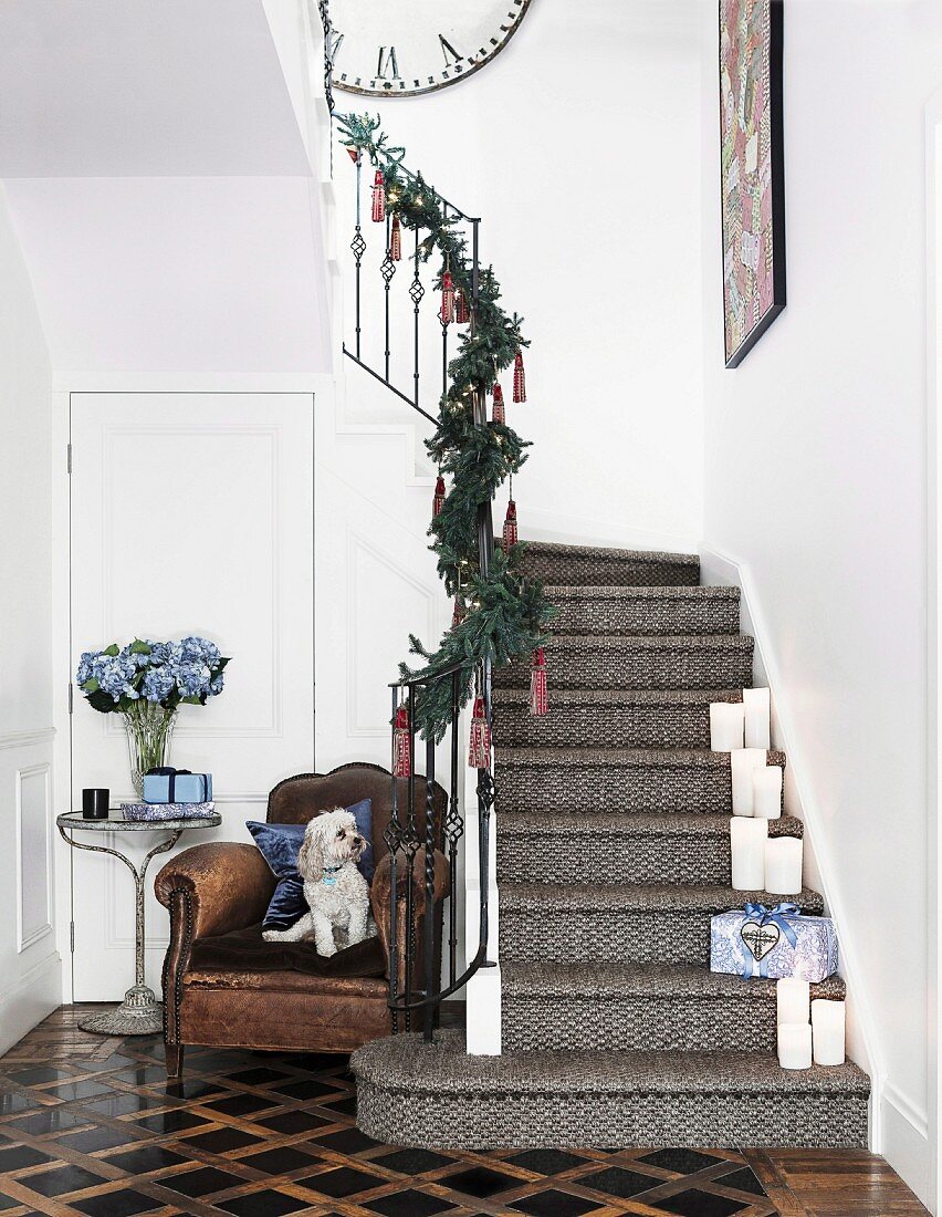 Staircase decorated for Christmas, white candles on steps, small dog on the side on a comfortable leather armchair