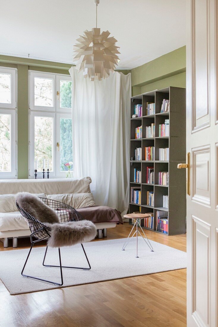 Klassikerstuhl mit Schaffell und Hocker vor Sofa am Fenster, in Zimmerecke Bücherregal und grüne Wandfarbe