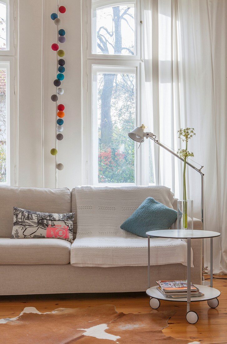 Runder, rollbarer Beistelltisch vor Sofa und Stehleuchte, an Fenster weißer Vorhang