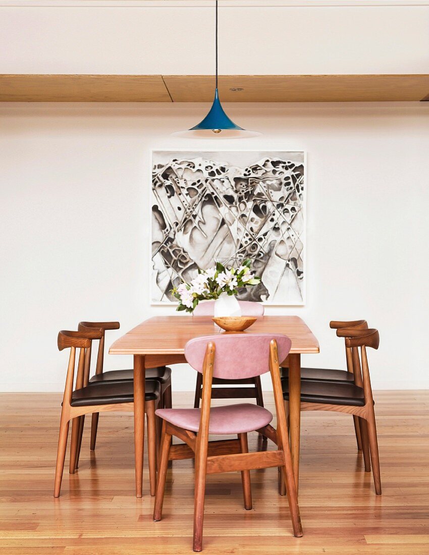 Holzstühle mit lederbezogenem Sitzpolster um Tisch mit Retroflair vor Wand mit modernem Bild