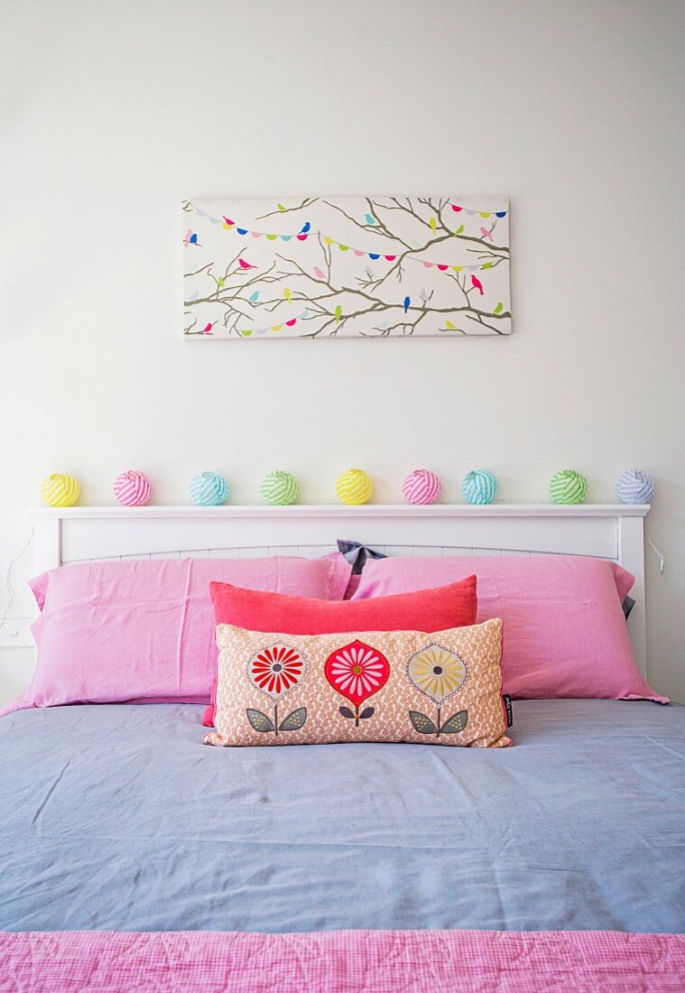 Mädchenzimmer mit rosafarbenen Kissenbezügen, pastellfarbener Lichterkette auf weißem Betthaupt und Vogelmotivbild an Wand