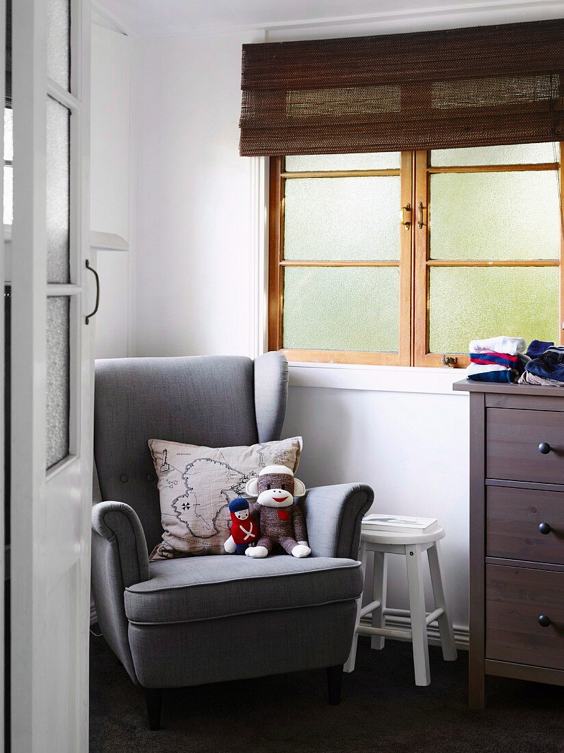 Blick durch offene Zimmertür auf grauen Lesesessel mit Stofftieren und Kissen, neben Sprossenfenster mit Bambusrollo im Kinderzimmer