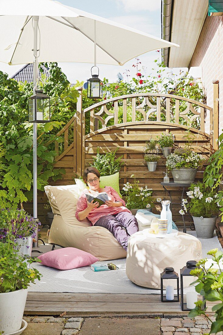 A woman relaxing under a parasol on a terrace with a wooden privacy fence with flower pots