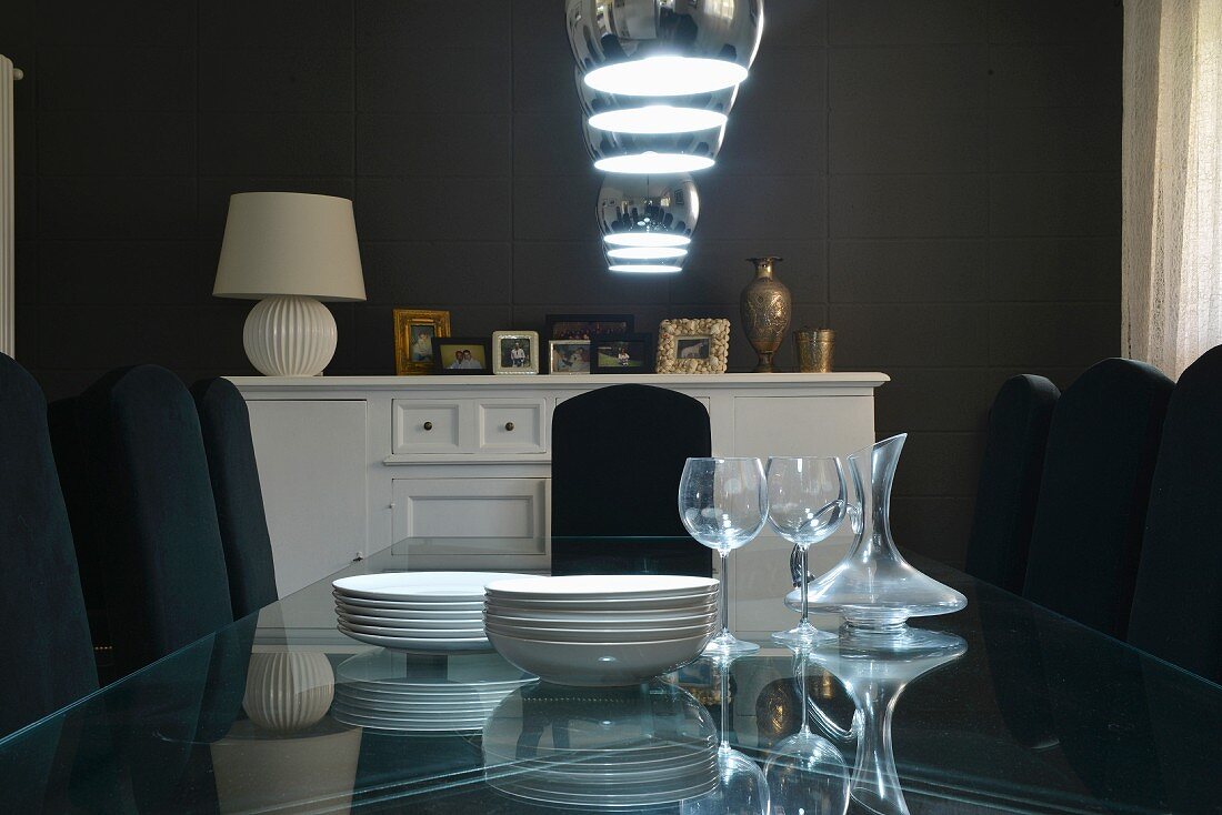 Crockery on glass table under pendant lamps in dining room with dark walls