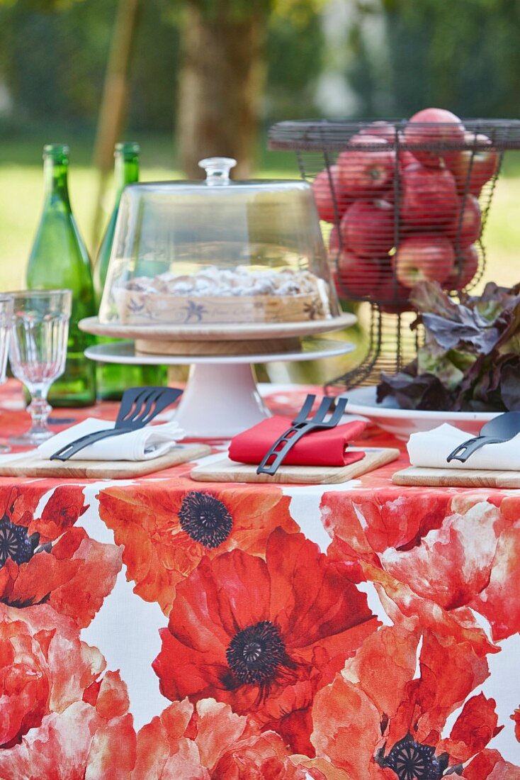 Tisch im Garten gedeckt mit roter Blumentischdecke, Kuchen und Korb mit Äpfeln