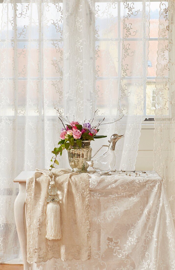 Flower arrangement and duck-shaped carafe on small table with off-white lace tablecloth in front of window with lace curtains