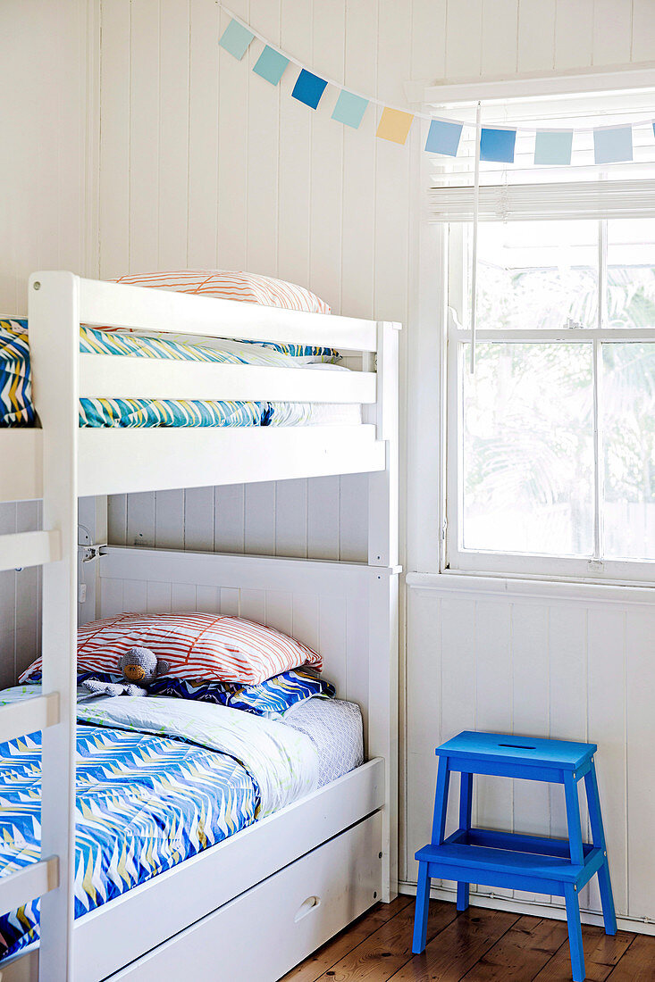 Weisses Stockbett mit bunter Bettwäsche und blauem Trittleiter-Hocker vor Fesnter
