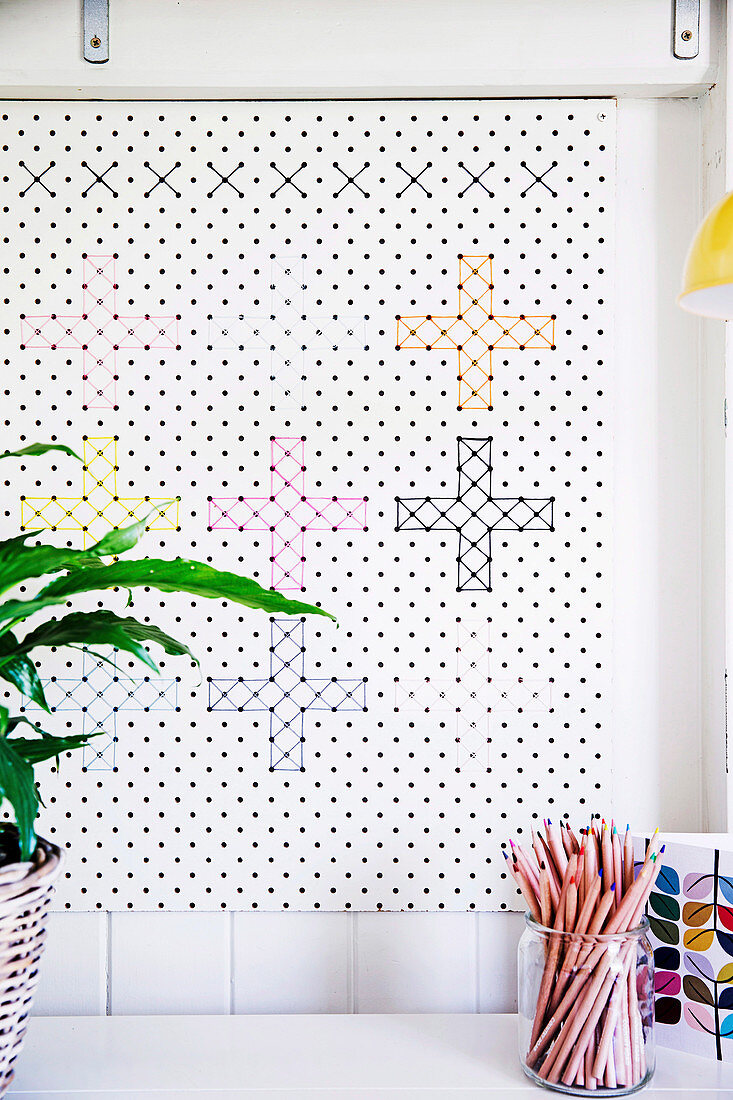 weiße Lochplatte mit Kreuzstichen in verschieden Farben als Wanddekoration, davor Glas mit Buntstiften
