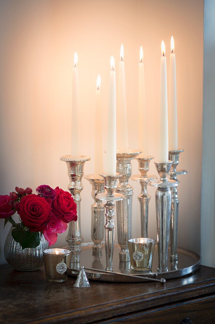 Lit candles in various silver candlesticks on tray and small vase of roses