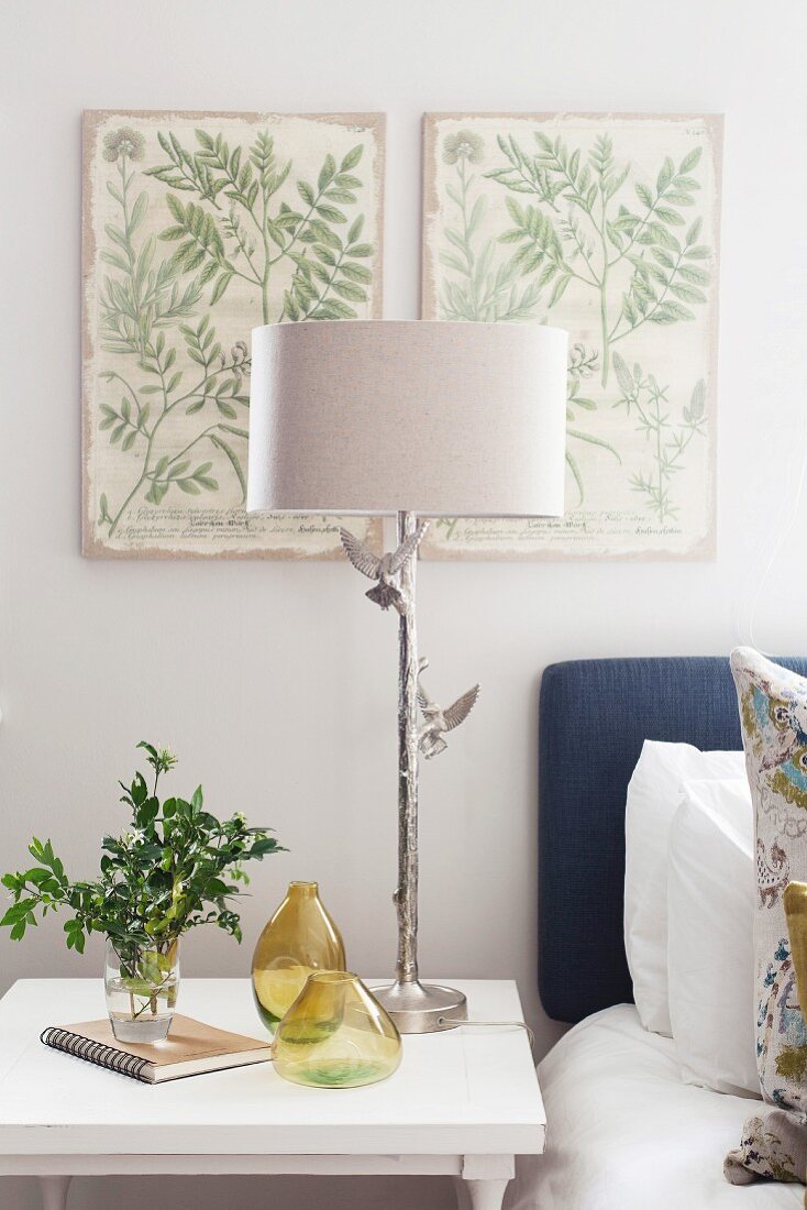 Bedside lamp with silver base and two bird ornaments below botanical illustrations on wall