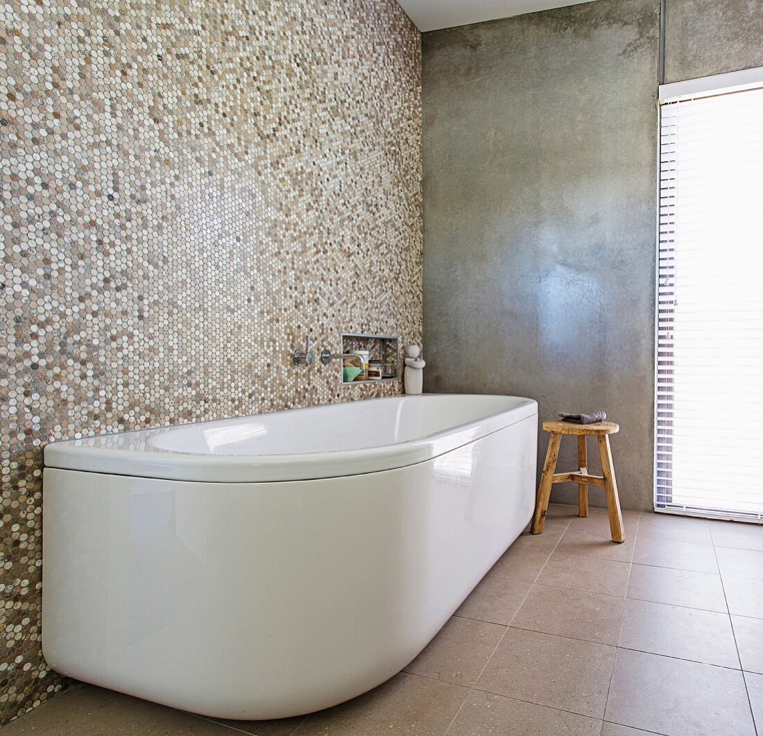 Freistehende, weiße Badewanne an Mosaikfliesenwand in minimalistischem Bad mit Betonwand
