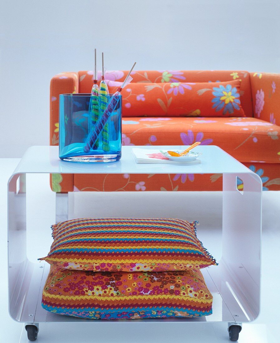 Colourful cushions with crocheted trim inside white metal coffee table in front of bright orange, floral sofa
