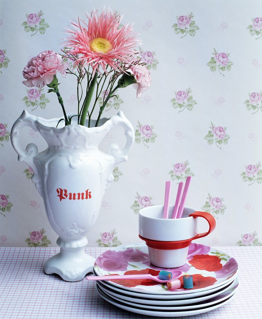 Stack of rose-patterned plates and cup next to flowers in white trophy vase against vintage-style rose-patterned wallpaper