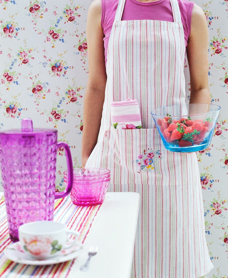 Woman wearing striped apron standing in front of floral wallpaper holding bowl of strawberries