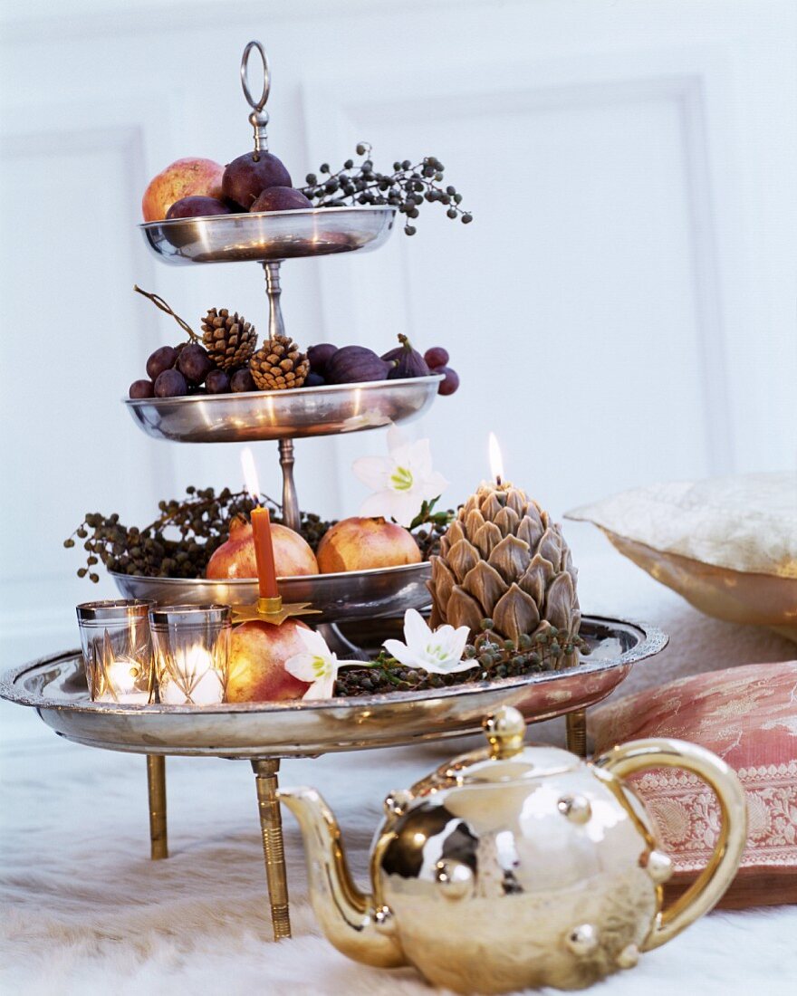 Silberglänzende Etagere mit Obst, Kunstblumen und Kerzen dekoriert, davor goldglänzende Teekanne