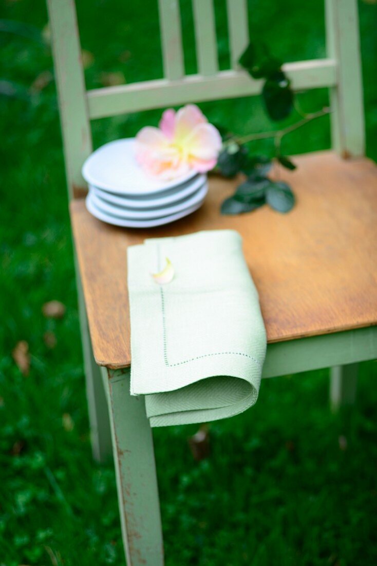 Stack of small plates, cut rose and linen napkin on chair seat