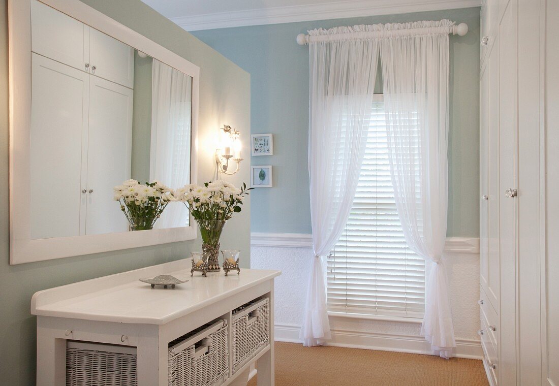 Console table with white wicker boxes below mirror on wall; draped curtains and louvre blinds on window in background