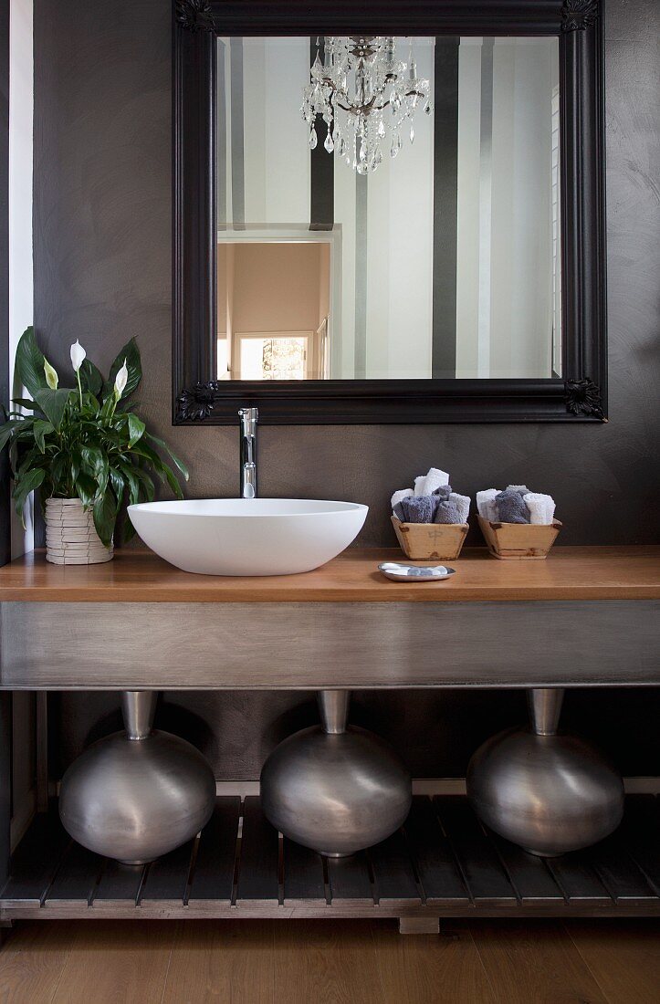 Modern washstand with white basin, three metal vases on shelf below and framed mirror on grey, marbled wall
