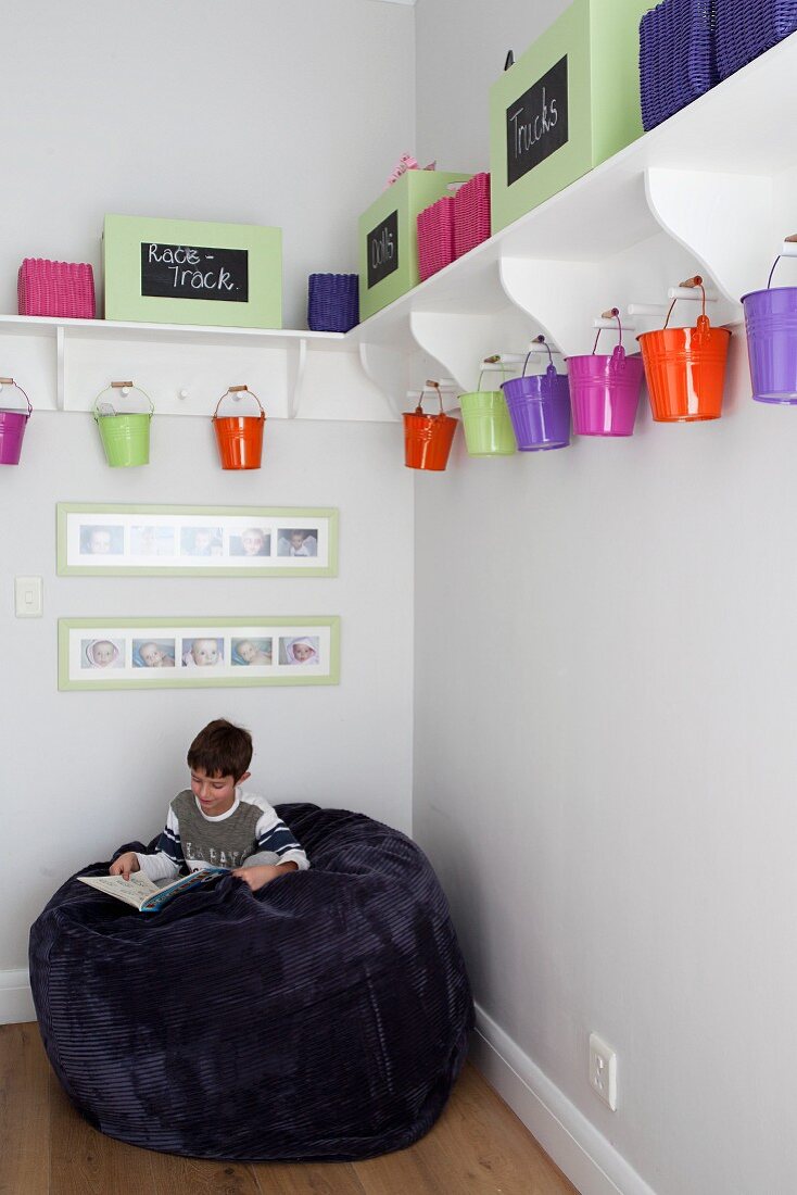 Kleiner Junge auf riesigem Sitzsack in Zimmerecke, oberhalb farbige Metalleimer an weisser Wandkonsole aufgehängt